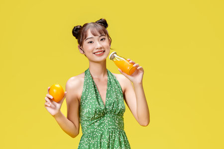 Excited asian woman drinking beverage freshness orange juice glass standing on yellow backgroundの写真素材
