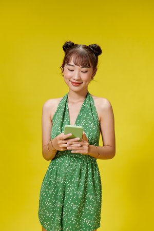 Beautiful Asian woman holding smartphone and smiling on light yellow backgroundの写真素材