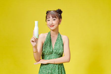 Portrait of beautiful woman holding bottle with cosmetics while standing isolated over yellow backgroundの写真素材