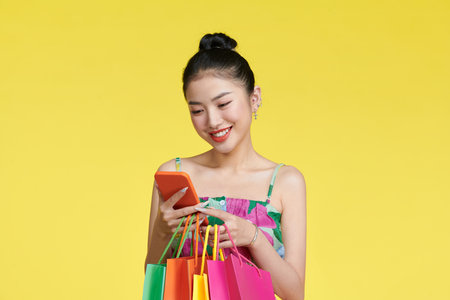 happy young asian woman celebrating with mobile phone and holding shopping bagsの写真素材