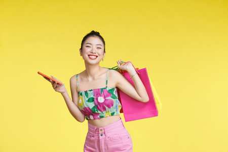 Young excited cheerful woman holding shopping bags isolated over yellow background using mobile phone looking aside.の写真素材