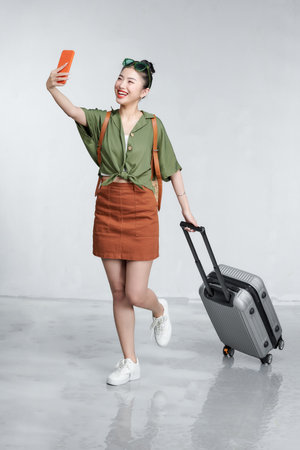Portrait of a cheerful beautiful young woman standing with a suitcase, holding mobile phoneの写真素材