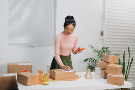 Smiling Asian online business owner prepares parcels and check online orders for delivery to customers on her smartphoneの写真素材