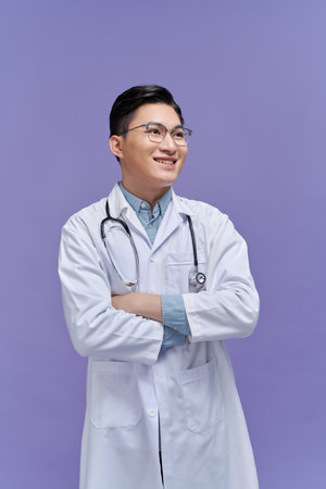 smiling young male doctor with stethoscope over neck in medical coat isolated on purpleの写真素材