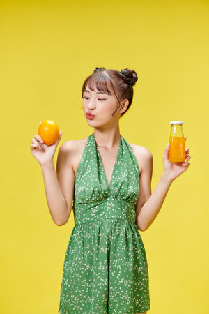 Tropical fruits season. Positive girl holding fresh orange and juice, yellow studio backgroundの写真素材