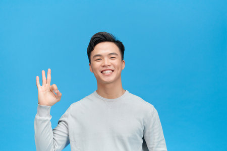 Portrait of handsome excited man smiling and showing ok sign at camera isolated over blue backgroundの写真素材