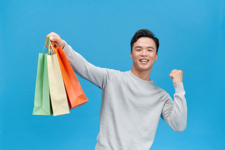 Smiling Asian man holding shopping bags raised fist up shout yeah isolated on blue backgroundの写真素材