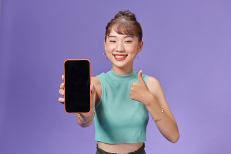 Portrait of smiling asian woman holding smartphone with black blank device screen in hand, showing thumb up gestureの写真素材