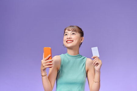 Asian happy young girl smiling cheerful and showing plastic credit card while holding mobile phoneの写真素材