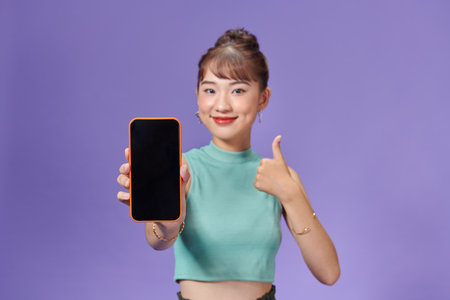 Satisfied happy asian girl show thumbs-up as demonstrate mobile phone screen applicationの写真素材