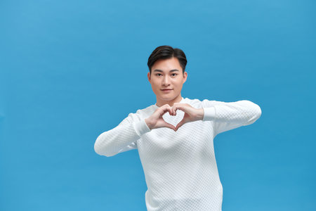 Portrait of attractive Asian man in red t-shirt speaks about own feelings, makes heart gesture over chest,の写真素材