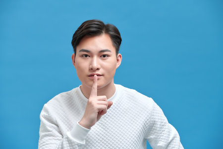 Portrait of serious young Asian man showing a sign of silence gesture putting fingers in mouthの写真素材