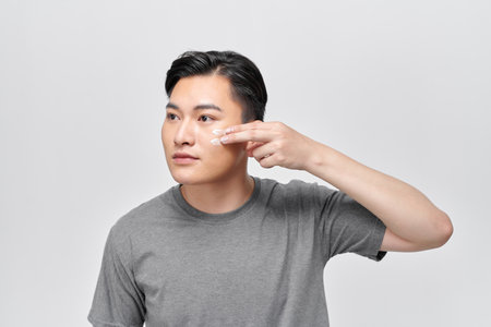 Portrait of handsome young man applying cream to his face, isolated on white backgroundの写真素材