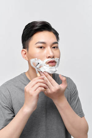 Positive attractive young guy with gel foam on lower face shaving with razor at mirrorの写真素材