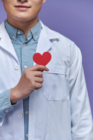 Young doctor with stethoscope over purple background holding red heartの写真素材