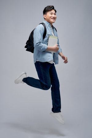 Full length of a smiling young asian man carrying backpack isolated, holding books, jumpingの写真素材