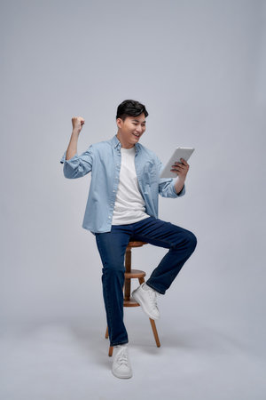 Cheerful casual man holding his tablet and celebrating with his fist in the air while sitting on a chairの写真素材