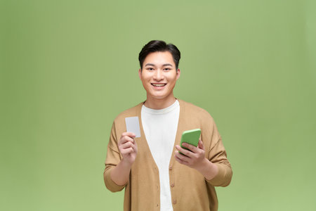 cheerful man keeping mobile phone showing bank credit card isolated on green background