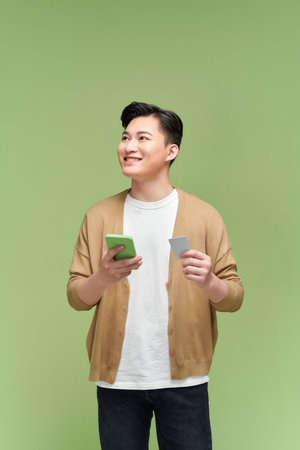 Smiling man holding credit card and mobile phone while standing against green backgroundの写真素材