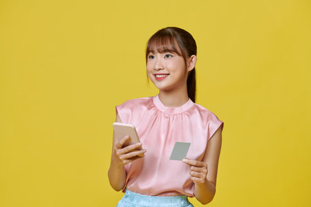 happy young asian woman using mobile phone and credit card isolated over yellow background.の写真素材