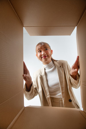 Cheerful Asian young woman opening cardboard box or parcel and looking insideの写真素材