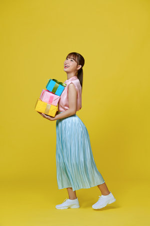 A smiling pretty girl holding stack of gift boxes isolated over yellow backgroundの写真素材
