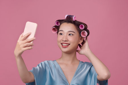 Happy young woman in dressed casual robe with hair curlers taking selfie on pink backgroundの写真素材