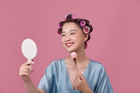 Cute girl with hair curlers doing makeup on her face at the makeup table, using makeup brushの写真素材