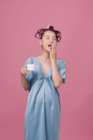 Happy young woman in dressing-gown and hair curlers drinking coffee on pink backgroundの写真素材