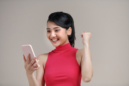 Happy young asian woman looking at screen of smartphone standing on isolated white background.の写真素材