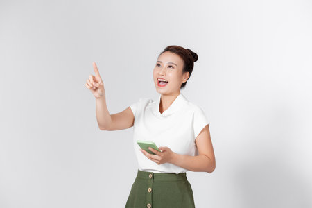 Overjoyed smiling happy cute Asian young lady point finger up hold phone posing isolated on white background.の写真素材