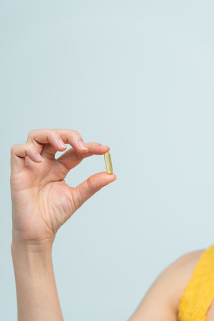 A close-up view of a hand holding a yellow fish oil capsule, emphasizing health and wellness concepts against a light background.の写真素材