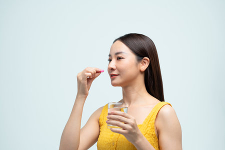 A smiling woman holding a glass of water and taking a pill while wearing a yellow dress.の写真素材