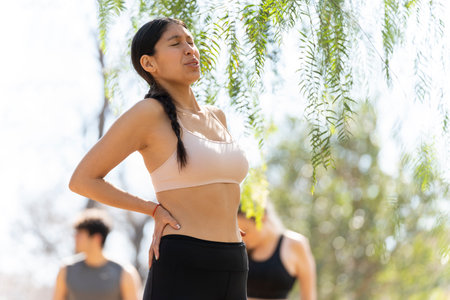 Hispanic young woman expresses back pain while doing sports in the parkの写真素材