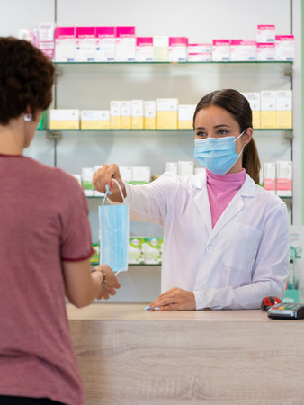A young female pharmacist in a mask giving another surgical mask to a customerの写真素材