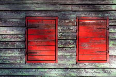Weathered vintage building facade with two closed shutters. Wooden wall with two windows, painted in red and green.の写真素材