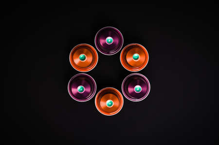 Less content, more packaging, higher price and even more waste. Arrangement of six aluminum coffee capsules in orange and purple against black background. Table top view, flat lay, copy space.の写真素材