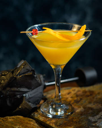 Martini glass filled with orange colored drink. 'Damn the weather' is a prohibition era cocktail based on gin, vermut, bitter orange liqueur and orange juice. Wet umbrella and dark background.の写真素材