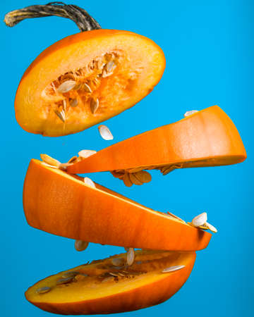 Small orange pumpkin sliced into multiple parts floating in the mid air. Blue Halloween decoration preparation. Food levitation photography. Squash seeds falling to the ground. Gravity denying food.の写真素材