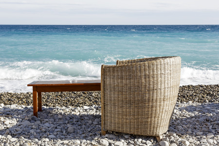 Wicker chair and wooden table on which ashtray stands on a rocky beach on the seafront 
