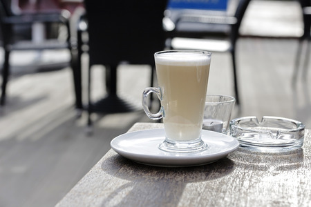 Tall cup filled with coffee with lots of milk stands on a wooden table, an empty glass ashtray is seen there as well.の写真素材