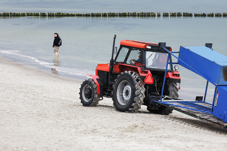 KOLOBRZEG, POLAND - JUNE 21, 2016: Tractor painted in red drive along the beach shore of the Baltic Sea, in the distance surprised by this fact the unidentified tourist can be seenのeditorial素材