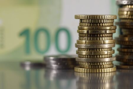 These are euro coins which have been stacked on a grey, flat surface and are visible against a blurred 100 euro banknote.の写真素材