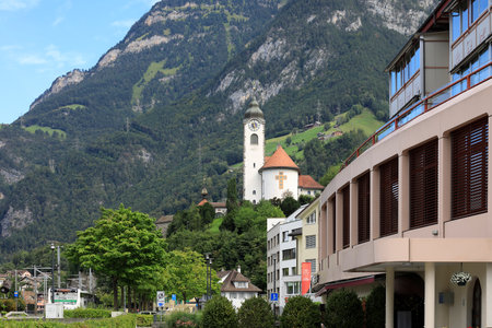 Fluelen, Switzerland - August 25, 2020: Town landscape with a church and a modern building. In the background is a steep, wooded mountain slope.のeditorial素材