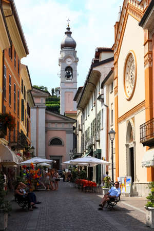 Menaggio, Italy - July 12, 2009: A bell tower is visible at the end of the narrow street. Between the buildings you can see stalls and tables under umbrellas.のeditorial素材