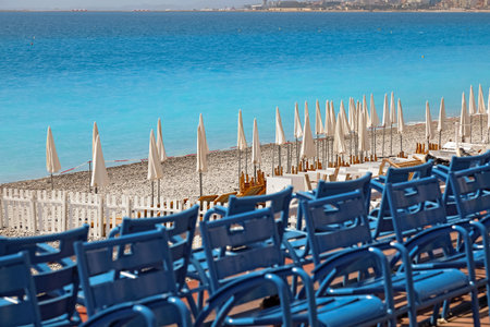 Beach umbrellas on a pebble beach for sunbathers to relax are not yet open. The famous blue chairs of Nice along the promenade are not yet occupied, they are all empty.の写真素材