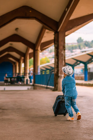 Little boy in a blue jacket and hat with a suitcase at the train stationの写真素材
