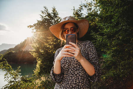woman with hat and sunglasses using mobile phone in sunset on the lake sideの写真素材
