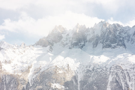 Snow-covered mountain range under a cloudy sky with sunlight breaking through, showcasing winter landscape and natural beautyの写真素材