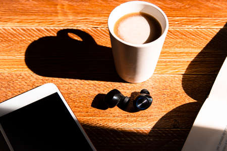 Zenital shot of a cup of coffee with earphones and a book on a sunlit wooden table. Concept of reading at home, home leisure, inner life and home relaxationの写真素材